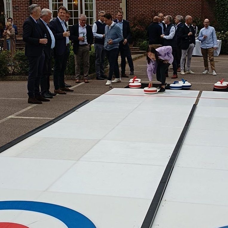 A group of office workers in suits watching someone trying out our hired in double lane Curling at an outdoor Corporate event.