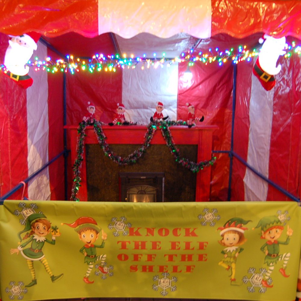 Christmas festive signage for classic Side Stall game throwing soft balls at cute little elves sat perched on a a bespoke handmade fireplace with a log burner that lights up and lots of tinsel, fairy lights and decorations.