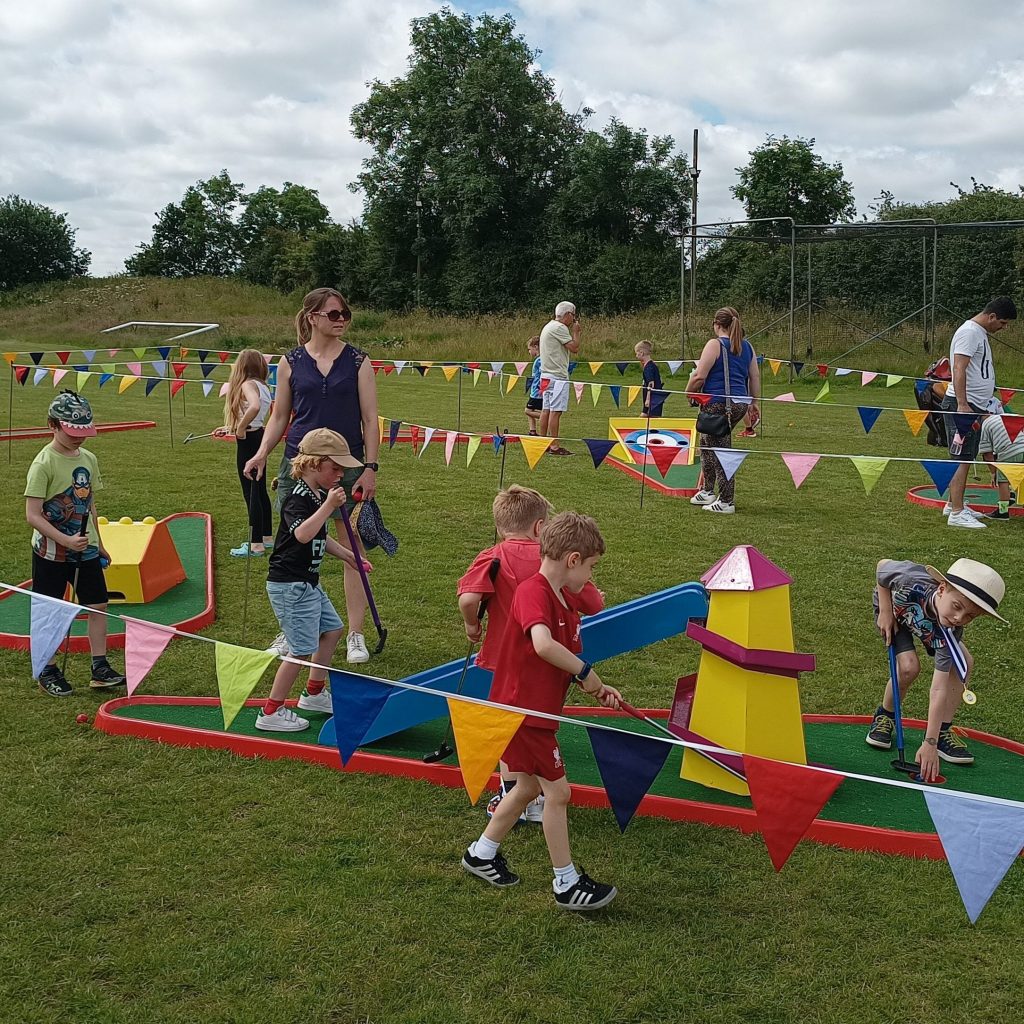 Lots of happy children with parents enjoying our Crazy Golf obstacles, with bright bunting flags all around.