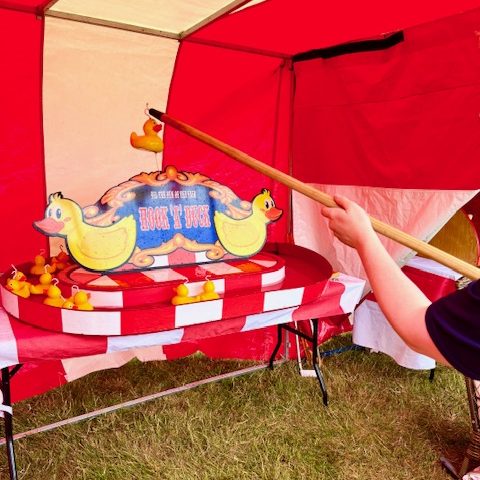 A man holding one of our Hook a Duck poles raised in the air to show the duck that has been successfully hooked, in our red and white Fete Game Side Stalls.