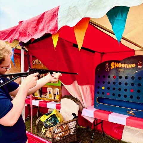 A man holding one of our cork shooting guns, aiming at our Shooting Gallery which is sat in our red and white Carnival Game Side Stalls.