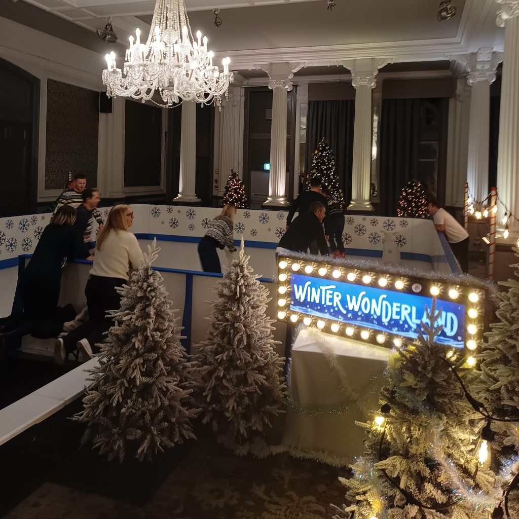 A custom snowflake decorated Synthetic Ice Rink with a Winter Wonderland sign surrounded by small snow covered artificial Christmas Trees all set up indoors with a stunning chandelier light above.