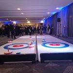 Two Twin Curling Lanes set up side by side indoors with a group of Corporate men and women at the far end taking turns.