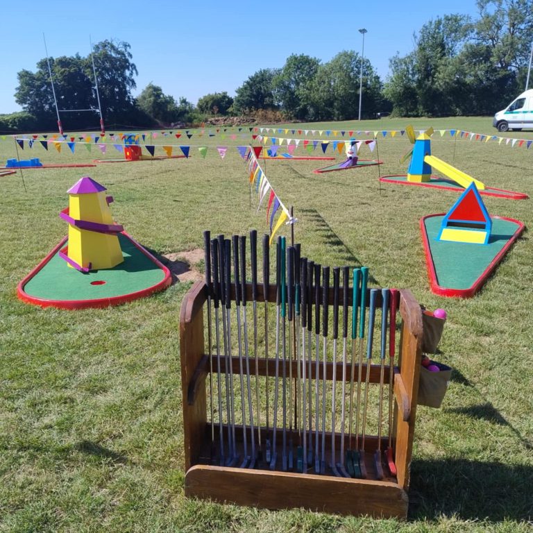 Our bespoke wooden Crazy Golf club and ball stand with various brightly painted obstacles behind sat on their red bases with artificial grass.