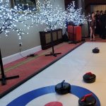 A Curling Lane indoors on carpet with light up festive trees on stands alongside and a group of office workers at the end enjoying the game.