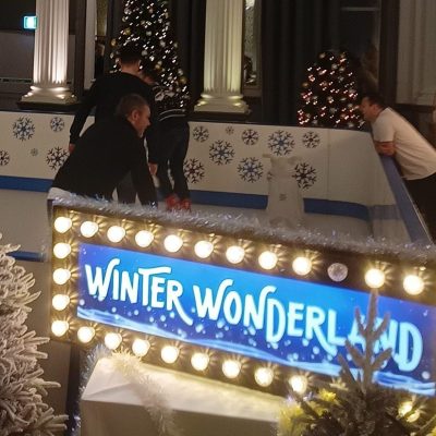 A custom snowflake decorated Synthetic Ice Rink with a Winter Wonderland sign surrounded by small snow covered artificial Christmas Trees all set up indoors with a stunning chandelier light above.